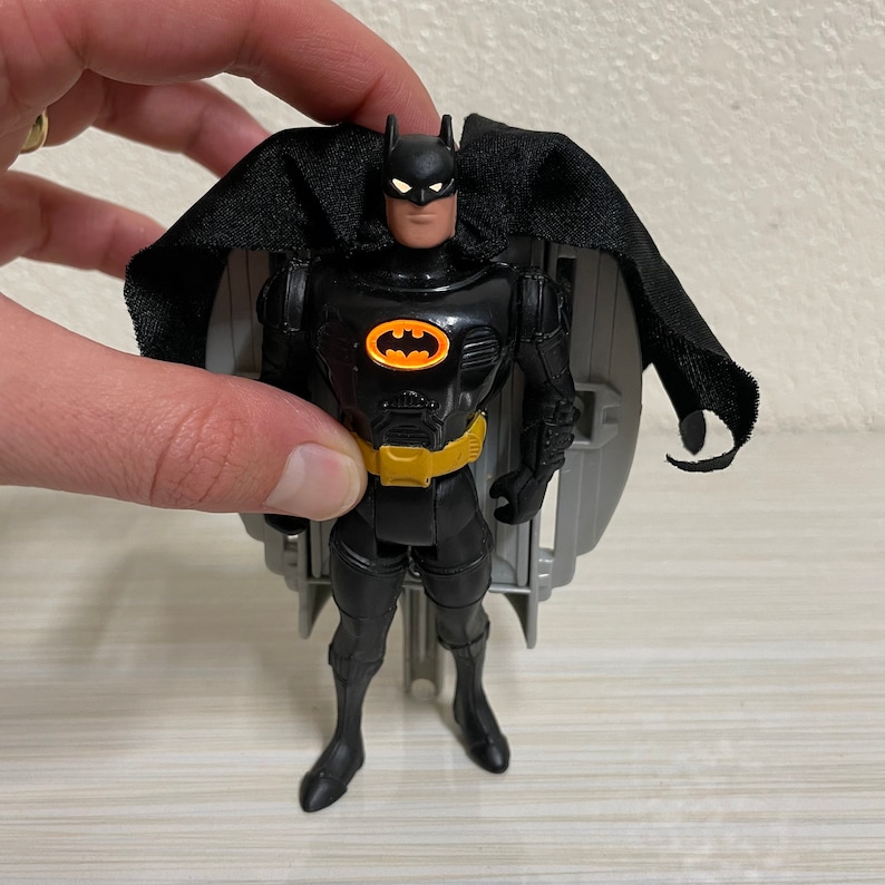 May include: Black Batman action figure with a black cape and a yellow utility belt. The figure is posed with its arms at its sides and its legs slightly apart. The figure is standing on a grey plastic base.