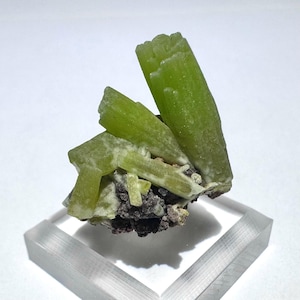 May include: A cluster of bright green crystals, possibly a mineral specimen, with a rough, textured surface. The crystals are arranged in a cluster on a dark, speckled base, presented on a clear, rectangular stand.