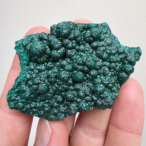 May include: A vibrant, dark green malachite specimen with a bubbly, textured surface. The mineral is held in a hand, showcasing its natural, organic form. The color is a rich, deep green, with a slightly rough texture.