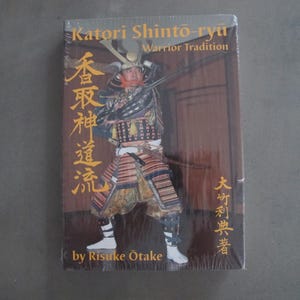 May include: A book titled "Katori Shinto-ryu Warrior Tradition" featuring a warrior in traditional Japanese armour. The cover has Japanese text and the author's name, Risuke Otake. The book is wrapped in clear plastic.
