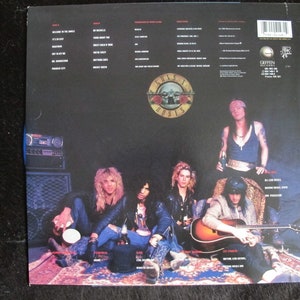 May include: The back cover of a Guns N' Roses vinyl record album. The album cover features a black and white photo of the band members sitting on a rug. The band members are wearing casual clothing. The album title "Guns N' Roses" is printed in white letters on a black background. The album track list is printed on the back cover.