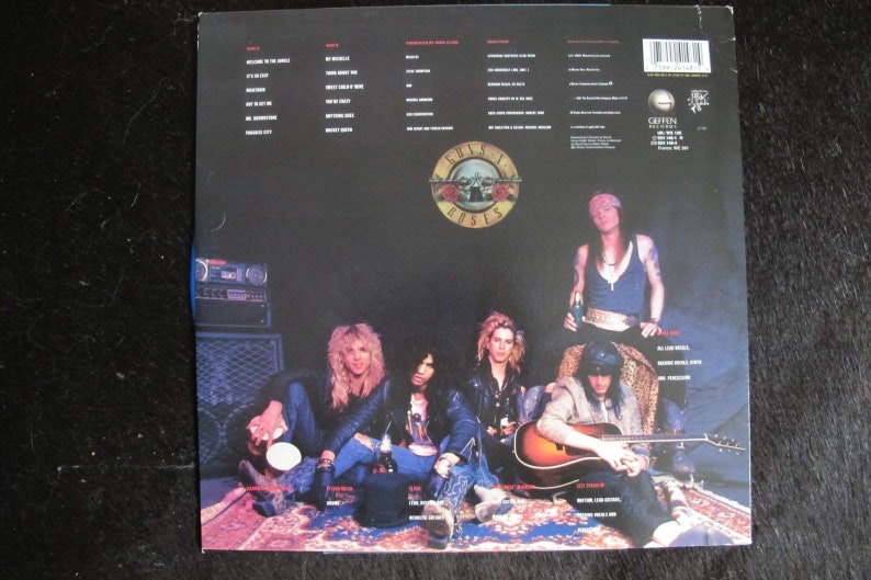 May include: The back cover of a Guns N' Roses vinyl record album. The album cover features a black and white photo of the band members sitting on a rug. The band members are wearing casual clothing. The album title "Guns N' Roses" is printed in white letters on a black background. The album track list is printed on the back cover.