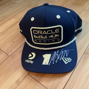May include: A navy blue baseball cap featuring an Oracle Red Bull Racing patch on the front, a gold braided cord across the brim, a gold number one, a Viaplay logo, a New Era logo, and a signature in silver ink on the curved brim of the cap.