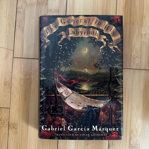 May include: A hardcover book titled "The General in His Labyrinth" by Gabriel García Márquez. The cover features a landscape with a hammock, trees, and a moonlit sky. The book is on a wooden surface.