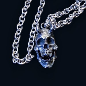 Op de afbeelding: Een zilveren ketting met een gedetailleerde schedelhanger met hoorns. De schedel heeft een dreigende uitdrukking met gebaarde tanden.