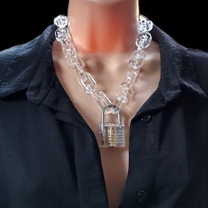 May include: Clear acrylic chain necklace with a silver padlock pendant. The necklace is made of large, transparent links. The padlock has a silver metal frame and a gold interior. The necklace is worn over a black shirt.