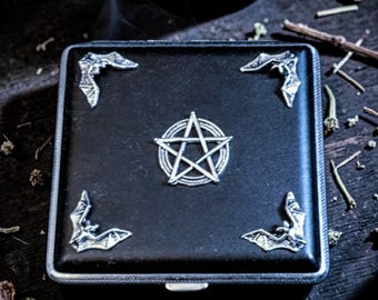 May include: Black square case with silver accents. The case features a silver pentagram in the center and silver bat designs in each corner. The case is on a dark wooden surface.