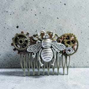 May include: A steampunk-style hair comb featuring a silver bee with detailed wings and body. The bee is flanked by bronze and copper-colored gears. The comb's teeth are a bronze color, creating a unique accessory.