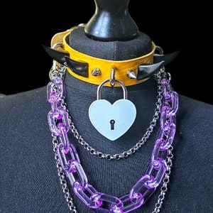 Op de afbeelding: Een gele leren choker met zwarte spikes en een zilveren hartvormig hangslot, gelaagd met een zilveren ketting en een paarse plastic ketting.