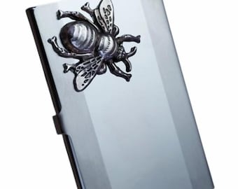 May include: A rectangular, metallic silver business card holder with a polished, reflective finish. Attached to the upper left corner is a three-dimensional, dark grey metal emblem shaped like a bee with detailed wings and body segments.
