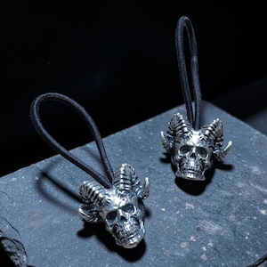 May include: Two black elastic hair ties, each featuring a metallic silver-toned skull charm with prominent curved horns. The skull charms have detailed facial features, including deep eye sockets and a defined jawline, attached to the black elastic bands.