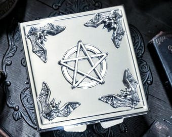 May include: Silver-colored square box with a pentagram design in the center and bat-shaped corner accents. The box is set on a dark, ornate wooden surface, with other dark-colored objects in the background.
