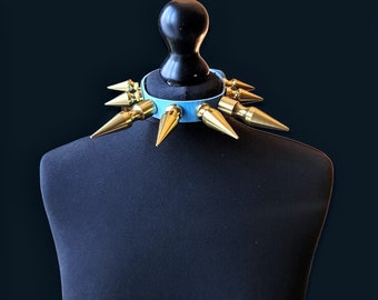 Op de afbeelding: Een lichtblauwe leren halsband met goudkleurige metalen spikes. De halsband wordt getoond op een zwarte mannequin. De spikes zijn gelijkmatig verdeeld rond de halsband, wat een gedurfde en edgy uitstraling creëert.