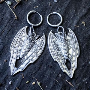 May include: A pair of silver bat earrings. Each earring has a detailed bat pendant with outstretched wings, suspended from a silver hoop. The bats have intricate wing patterns, set against a dark, textured background.