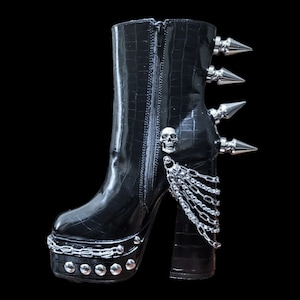 Pode incluir: Botas de plataforma de couro preto com um amuleto de caveira, correntes prateadas e pontas prateadas no calcanhar.