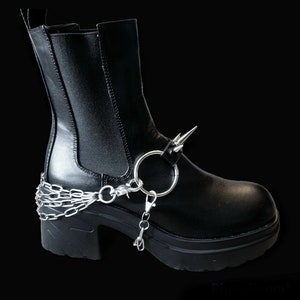 Puede incluir: Botas negras de tobillo con tacón grueso y detalle de cadena y anillo plateado. Las botas tienen punta puntiaguda y una espiga plateada en el lateral.