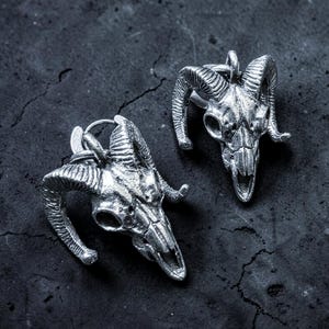 May include: Two silver-colored ram skull pendants with detailed horns and facial features. The pendants are displayed on a dark, textured surface, showcasing their gothic and edgy design. The skulls are facing different directions.
