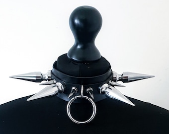 May include: A black collar featuring multiple large, silver-toned metal spikes arranged around the band. A circular metal ring is attached to the front center, positioned between the spikes. The collar is displayed on a black mannequin neck form.