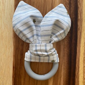 May include: A baby teether with a gray silicone ring and a fabric top shaped like bunny ears. The fabric is light blue and white striped with small bee illustrations.