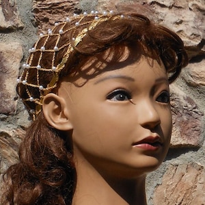 Op de afbeelding: Een gouden en witte parel haarband met een delicaat kettingontwerp. De haarband wordt gedragen door een mannequin met bruin haar.