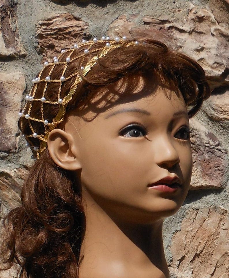 Op de afbeelding: Een gouden en witte parel haarband met een delicaat kettingontwerp. De haarband wordt gedragen door een mannequin met bruin haar.