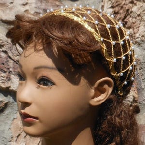 Op de afbeelding: Een gouden en witte haar kam met een net ontwerp en parel accenten. De kam wordt gedragen door een mannequin met bruin haar.