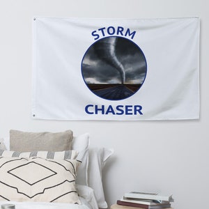 May include: White flag with the words "STORM CHASER" in blue, with a circular graphic of a tornado over a road. The flag is hanging on a white wall above a bed with decorative pillows and a wooden side table.