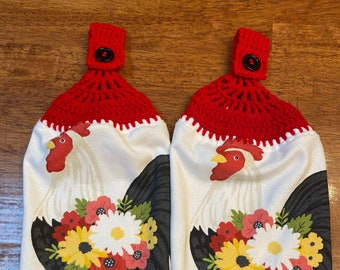Op de afbeelding: Twee witte keukendoeken met een hanenontwerp. De hanen hebben zwarte vleugels, rode kammen en bloemboeketten. De handdoeken hebben een rode gehaakte bovenkant met een zwarte knoop om op te hangen.