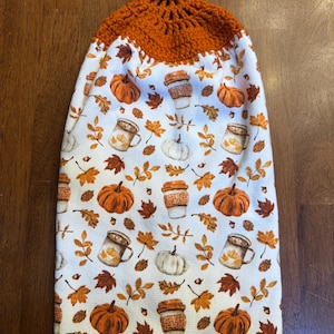 May include: A white kitchen towel with an orange crochet top, featuring a fall-themed pattern. The pattern includes orange pumpkins, coffee cups, and autumn leaves. The towel has a button closure and a decorative orange pom-pom trim.