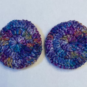 May include: Two handmade, circular coasters. Each coaster is crocheted with a thick, multicolored yarn. The yarn features shades of blue, purple, and a touch of yellow and orange. The coasters are designed with a spiral pattern.