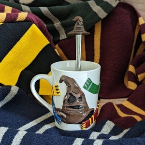 Puede incluir: Una taza de cerámica blanca con una imagen esculpida en 3D del Sombrero Seleccionador de la serie Harry Potter. La taza está rodeada de bufandas en los colores de las casas de Hogwarts: Gryffindor, Slytherin, Hufflepuff y Ravenclaw.