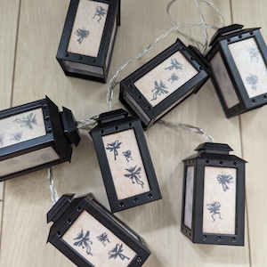 Könnte beinhalten: Eine Lichterkette mit schwarzen, laternenförmigen Glühbirnen. Jede Glühbirne hat einen weißen Hintergrund mit einer Silhouette einer Fee darauf.