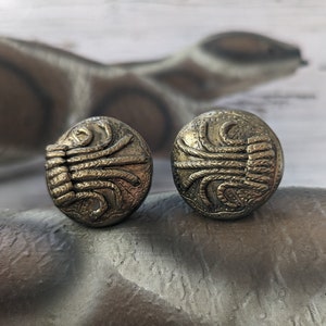 May include: A pair of gold-toned earrings with a geometric design. The earrings are round and have a textured surface.