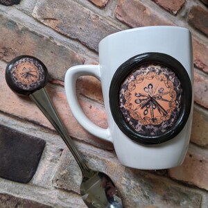 May include: A white ceramic mug with a black circular design and a matching spoon. The design on the mug and spoon features an orange and brown circular pattern with text. The spoon is stainless steel.