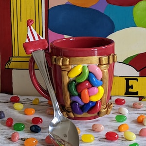 May include: A red ceramic mug with a gold rim and a jelly bean design. The mug has a spoon with a red and white striped hat on the end. The mug is surrounded by colorful jelly beans.