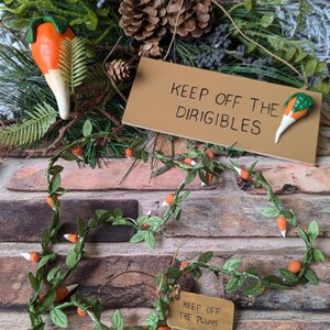 May include: A set of two brown wooden signs with the text "Keep off the Dirigibles" and "Keep off the Plums". The signs are decorated with orange and white carrot-shaped ornaments. A garland of green leaves and orange and white carrot-shaped ornaments is also included.