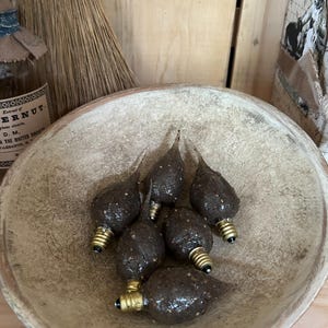 May include: A collection of brown, teardrop-shaped light bulbs with gold-colored bases rests in a rustic, wooden bowl. The bulbs have a textured surface and are arranged in a cluster. The bowl is light brown and has a worn appearance.
