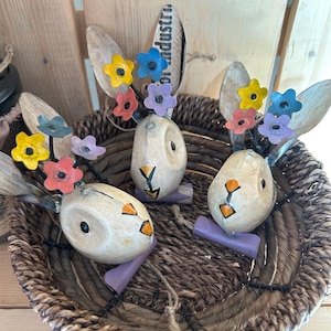 May include: Three Easter bunny figurines with spoon ears and colorful flower crowns sit in a woven basket. The bunnies have painted faces and purple bases. The basket is on a wooden surface.