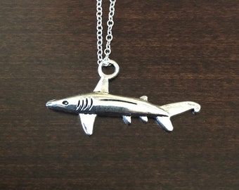May include: A silver shark pendant necklace on a silver chain. The shark is facing to the right and has a slightly curved tail.
