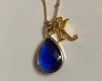 May include: A gold-colored necklace featuring a teardrop-shaped, faceted, deep blue pendant. A gold letter "K" charm hangs next to the pendant. The necklace has a delicate chain with small, evenly spaced beads.