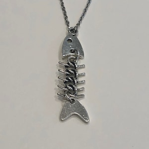 May include: A silver-colored fish skeleton pendant necklace. The pendant is a detailed representation of a fish skeleton, with a chain connecting the pendant to the necklace. The necklace is displayed against a white background.