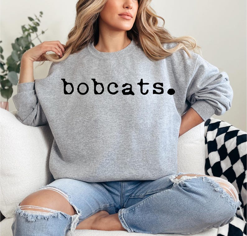 May include: A heather gray sweatshirt with the word "bobcats." printed in black. The sweatshirt is a crew neck style and is paired with light blue jeans with distressed details.