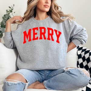 May include: A gray sweatshirt with the word "MERRY" printed in bold red letters. The sweatshirt is a crew neck style and is being worn with ripped blue jeans. The overall theme is casual holiday apparel.