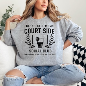 May include: A light gray sweatshirt with the text "BASKETBALL MOMS COURT SIDE SOCIAL CLUB" in black. A basketball hoop graphic is centered below the text. The bottom of the sweatshirt reads "WARNING: MAY YELL AT THE REF".