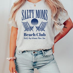 以下が含まれることがあります： 青い文字で「Salty Moms Beach Club Girls Just Wanna Have Sun」と書かれたグレーのTシャツ。デザインにはビーチチェア2つと波のある太陽が含まれています。
