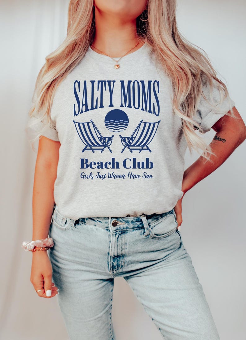 以下が含まれることがあります： 青い文字で「Salty Moms Beach Club Girls Just Wanna Have Sun」と書かれたグレーのTシャツ。デザインにはビーチチェア2つと波のある太陽が含まれています。