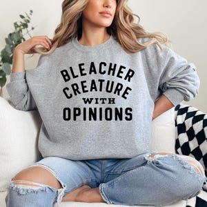Puede incluir: Una sudadera gris con el texto negro "BLEACHER CREATURE WITH OPINIONS". La sudadera de manga larga tiene cuello redondo. La persona lleva vaqueros azules rotos.
