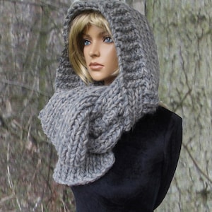 Könnte beinhalten: Ein grauer gestrickter Kapuzen-Schal mit einer grobmaschigen Strickstruktur. Der Schal wird von einem Model mit blonden Haaren getragen.