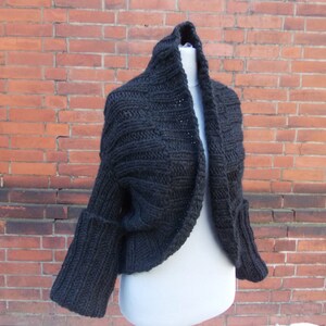Könnte beinhalten: Ein schwarzer, gestrickter, verkürzter Cardigan mit langen Ärmeln und einem weiten, offenen Kragen.