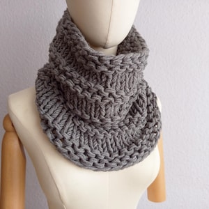 Può includere: Sciarpa a cerchio grigia a maglia grossa, esposta su un manichino. La sciarpa è realizzata con filato spesso, creando un aspetto strutturato. Il colore neutro la rende un accessorio versatile. La sciarpa è progettata per essere indossata intorno al collo.